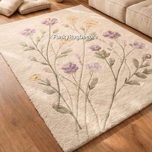 May include: Cream-colored floral area rug with a botanical design. The rug features purple and yellow flowers with green stems and leaves. The rug is rectangular and placed on a hardwood floor.