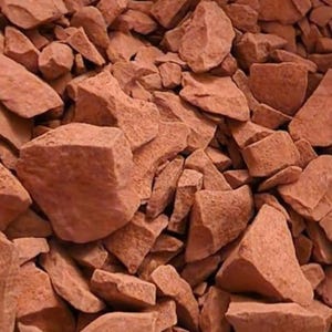 May include: A close-up shot of a pile of broken, reddish-brown rocks. The rocks vary in size and shape, with rough, uneven surfaces. The lighting creates shadows, adding depth and texture to the composition. The overall impression is of a natural, earthy material.