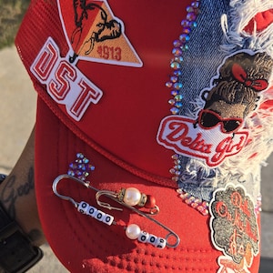 May include: A red trucker hat featuring a mesh back, a denim patch, and various embellishments including rhinestones, embroidered patches with the text DST, Delta Girl, and 1913, and two large safety pins decorated with white beads and pearls.