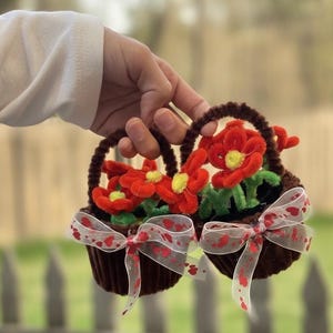 Puede incluir: Dos pequeñas cestas marrones llenas de flores rojas de limpiapipas con centros amarillos y hojas verdes. Cada cesta tiene un asa marrón y está adornada con una cinta blanca decorada con corazones rojos.