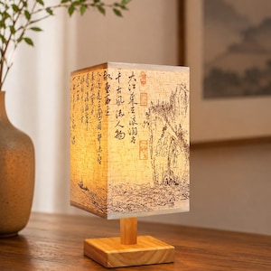 May include: A rectangular table lamp with a light-colored shade featuring black ink-style illustrations of mountains, water, and Chinese calligraphy. The lamp has a simple wooden base and a short cylindrical wooden stem supporting the shade.