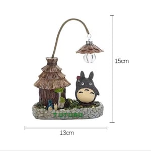 May include: A decorative lamp featuring a Totoro figure, a small house, and a lamp post. The lamp is approximately 5.9 inches tall and 5.1 inches wide. The base is a stone-like texture, and the lamp post has a woven lampshade.