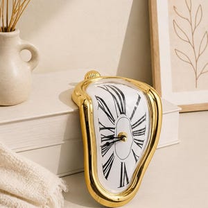 May include: A gold-colored, melting clock with a white face and black Roman numerals. The clock has a classic design with a modern twist, resembling a surrealist art piece. The clock is displayed on a white surface.