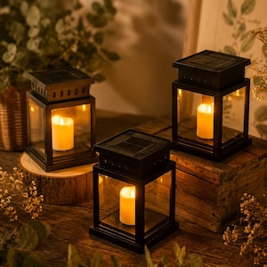 May include: Three black metal lanterns with glass panels and a lit candle inside. The lanterns are square-shaped with a solar panel on top, sitting on a wooden surface. The scene is warmly lit with a rustic, cozy feel.