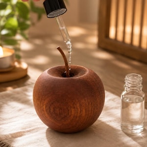 May include: A round, dark brown wooden apple-shaped diffuser with a small stem. A clear liquid is being dispensed from a black-capped glass dropper into the center of the wooden apple. A small, clear glass bottle sits next to the wooden object.