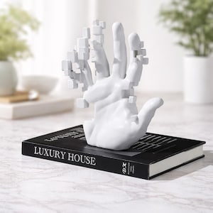 May include: A white ceramic hand sculpture with a pixelated design, resting on a black book titled "LUXURY HOUSE". The book is on a marble surface, with plants in the background.