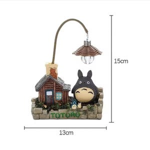 May include: A decorative lamp featuring a miniature scene with a house, a Totoro figure, and a small lamp. The lamp is 15cm tall and 13cm wide. The base has the word "TOTORO" in green. The lamp has a brown woven lampshade.