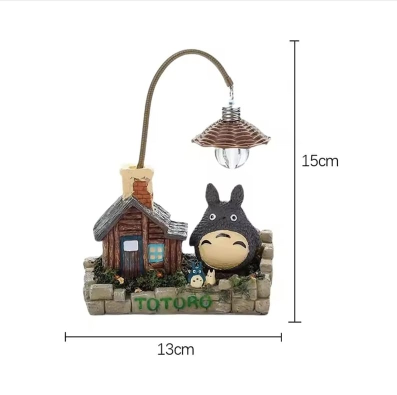 May include: A decorative lamp featuring a miniature scene with a house, a Totoro figure, and a small lamp. The lamp is 15cm tall and 13cm wide. The base has the word "TOTORO" in green. The lamp has a brown woven lampshade.