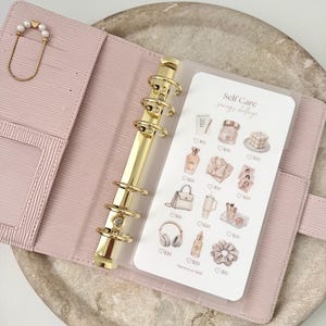 Peut inclure: Un classeur à six anneaux rose clair avec un mécanisme en métal doré et un trombone orné de perles. À l'intérieur se trouve une carte blanche intitulée Self Care Savings Challenge avec des illustrations d'articles de beauté et accessoires.