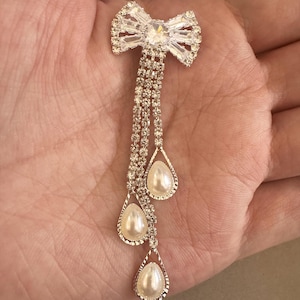 Op de afbeelding: Een zilverkleurige broche in de vorm van een strik, versierd met heldere kristallen accenten en drie hangende druppelvormige hangers, elk met een imitatieparel. De broche heeft meerdere strengen kleine kristallen.