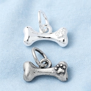 May include: Two dog bone charms with attached jump rings. The top charm has a polished, shiny silver finish, while the bottom charm features an oxidized, matte silver finish. Each bone-shaped charm is embossed with a small paw print design.