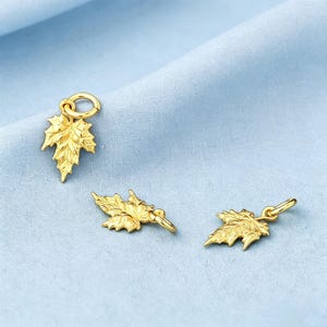 May include: Three gold-colored metal charms shaped like maple leaves, each featuring a textured surface and a small circular jump ring attached to the top for hanging. The charms are arranged in a scattered formation, displaying detailed veins.
