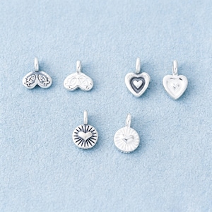 May include: Six small silver-toned metal charms with loops for hanging. The charms feature various embossed designs including two fish, a heart within a heart, a plain heart, and two circular charms with a central heart surrounded by radiating lines.