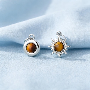 May include: Two silver-toned metal charms featuring round, polished tiger's eye gemstones. One charm is a simple circular disc, while the other is shaped like a sun with radiating points. Each charm includes a small circular jump ring for attachment.