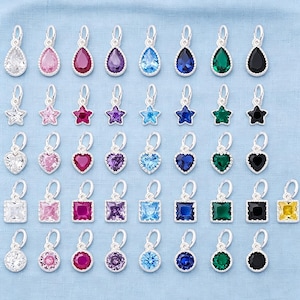 Puede incluir: Una colección de dijes de metal plateado con piedras preciosas facetadas en varias formas, incluyendo gotas, estrellas, corazones, cuadrados y círculos. Los dijes están dispuestos en filas, mostrando un espectro de colores como transparente, rosa, rojo, morado, azul, verde, negro y amarillo.