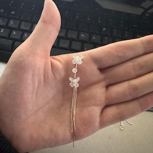 May include: A delicate silver earring with a floral and butterfly design. The earring features two flower-shaped and one butterfly-shaped crystal embellishments, connected by a thin chain with dangling strands.