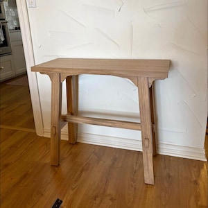Peut inclure: Une table d'appoint en bois brun clair avec un plateau rectangulaire et quatre pieds. La table a un design simple et minimaliste avec un grain de bois visible. Les pieds sont reliés par une barre de support horizontale. La table mesure environ 76 cm de long.