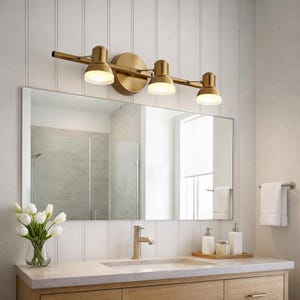 May include: A brass-finished three-light vanity fixture. The adjustable lights are mounted on a horizontal bar, positioned above a rectangular mirror. The bathroom features a light wood vanity and white walls. The mirror reflects the bathroom's interior.