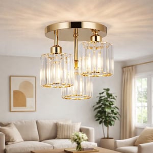 May include: A gold ceiling light with three clear glass shades. The shades are cylindrical with vertical lines, and the fixture has a circular base. The light is on, illuminating the room. The fixture is in a living room setting.