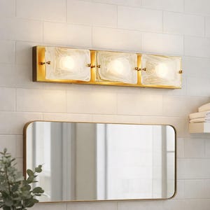 May include: A gold-toned vanity light fixture with three square, frosted glass shades. The fixture is mounted above a rectangular mirror with rounded corners. The wall is tiled in white.