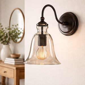 Puede incluir: Una lámpara de pared con una pantalla de vidrio transparente y un acabado en bronce oscuro. La lámpara tiene un brazo curvo y una placa trasera redonda. La bombilla es visible dentro de la pantalla de vidrio.