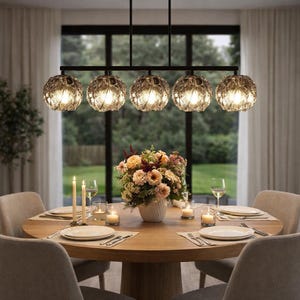 May include: A dining room setting with a round wooden table set for a meal. A floral arrangement sits in the center, with candles and place settings around the table. Above, a black chandelier with five globe-shaped lights illuminates the scene.