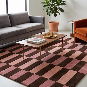 May include: A living room scene featuring a brown and pink checkered rug, a gray sofa, and an orange armchair. A wooden coffee table with gold legs holds a book and a tray with a candle. A potted plant adds a touch of greenery.
