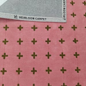 Peut inclure: Un tapis rose avec un motif répétitif de signes plus vert olive foncé disposés en grille. Le bord supérieur montre un envers gris avec une étiquette en tissu blanc imprimée du texte HEIRLOOM CARPET et un logo avec les lettres H et C.