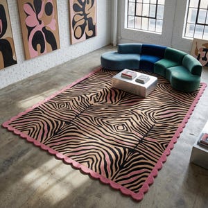 Puede incluir: Una alfombra rectangular con un patrón de rayas negras y beige similar al de una cebra y un borde festoneado de color rosa. La alfombra está colocada sobre un suelo de hormigón en una habitación con un sofá curvo azul y verde y una mesa.