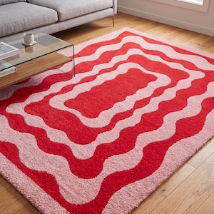May include: A rectangular rug with a wavy pattern in shades of red and pink. The rug is placed on a hardwood floor, and a gray sofa and glass coffee table are visible in the background. The rug's design creates a bold, eye-catching statement.