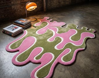 Könnte beinhalten: Abstrakter Teppich mit fließendem Design in Rosa und Olivgrün, umrandet mit Weiß. Der Teppich liegt auf einem Betonboden. Eine Retro-Orange-Lampe und Bücher sind im Hintergrund zu sehen. Das Design erinnert an eine flüssige oder organische Form.