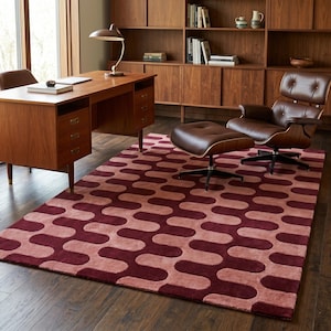 May include: A rectangular area rug featuring a repeating pattern of interlocking wavy shapes in shades of deep burgundy and soft dusty pink. The rug is positioned on a dark wood floor in a room with a wooden desk, a lounge chair, and a bookshelf.