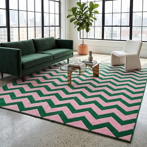 May include: A green velvet sofa and a white chair sit on a pink and green zigzag patterned rug. A glass coffee table with gold legs holds books and a vase. A potted plant is in the background.