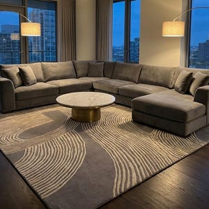 Op de afbeelding: Een moderne woonkamer met een grote grijze hoekbank, een ronde marmeren salontafel met een gouden voet en een vloerkleed met patroon. Twee gebogen vloerlampen met beige tinten zorgen voor sfeerverlichting. De kamer heeft grote ramen met uitzicht op de stad.