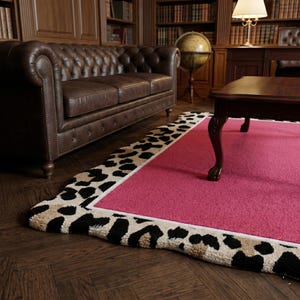 May include: A vibrant pink rug with a leopard print border. The rug is in a room with a brown leather sofa, a dark wood coffee table, and a bookcase. The rug's border features black and tan spots on a cream background.