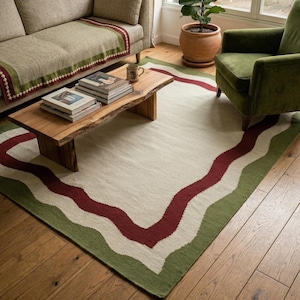 Puede incluir: Una alfombra rectangular con un centro color crema y un diseño de borde ondulado. El borde presenta una banda de color rojo oscuro y una banda verde. La alfombra está colocada sobre un suelo de madera, en un salón.