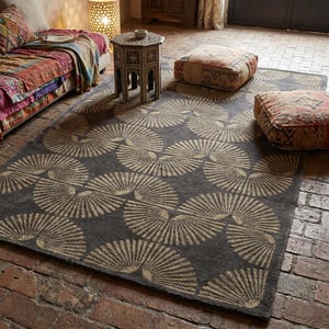 May include: A rectangular area rug featuring a dark gray background with a repeating pattern of light beige fan-shaped motifs. The rug is laid out on a brick floor, positioned near a sofa with a colorful throw, a small wooden side table, and two square floor cushions.