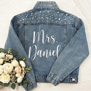 May include: A light blue denim jacket with pearl embellishments on the shoulders and the text "Mrs. Daniel" in white script on the back. The date "11.20.2026" is embroidered on the collar. A bridal bouquet with white and pink flowers is in the foreground.