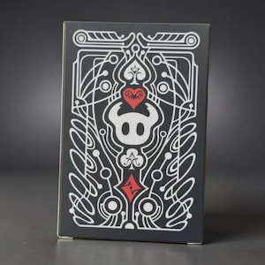 May include: A black playing card box featuring a white and red design. The design includes a heart, a spade, a diamond, and a clover. The box has a symmetrical pattern with a central character.