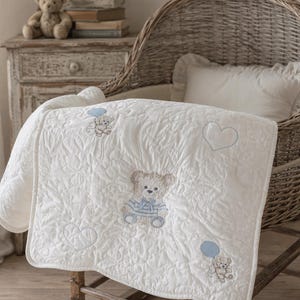 May include: A cream-coloured quilted baby blanket featuring embroidered teddy bears wearing blue striped shirts, light blue balloons, and heart outlines. The fabric has a textured, embossed floral pattern throughout the entire surface.