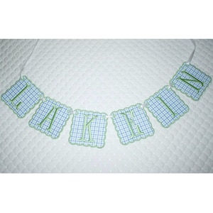 May include: A banner spelling out the word "THANK YOU" in green embroidered letters. Each letter is on a blue and white checkered flag with a scalloped green border. The banner is strung on a white ribbon.