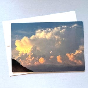 May include: A rectangular greeting card featuring a photograph of a dramatic cloud formation with golden and white hues against a blue sky. A white envelope is behind the card. The card's image includes a dark, mountainous silhouette.