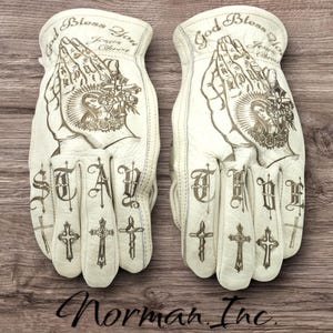 May include: A pair of off-white leather work gloves. The gloves feature detailed illustrations of praying hands, crosses, and the words "God Bless You Jesus Christ" and "Stay True". The gloves are branded with "Norman Inc."