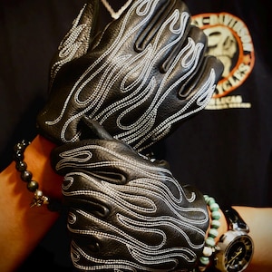 May include: Black leather gloves featuring white stitched flame patterns across the back of the hands. The gloves have a textured surface and are designed to cover the fingers and palms, showcasing detailed embroidery in a flame motif.