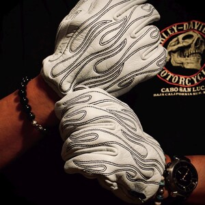 May include: A pair of white leather gloves featuring a black stitched flame pattern across the back of the hands. The gloves have a textured surface and are designed to cover the fingers and palms, showcasing detailed embroidery in a flame motif.