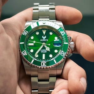 Puede incluir: Un reloj de pulsera de acero inoxidable con esfera y bisel verdes. La esfera del reloj muestra la marca "AURYX" y el texto "BYSSNA 300M AUTOMATIC". El reloj se sostiene en una mano.