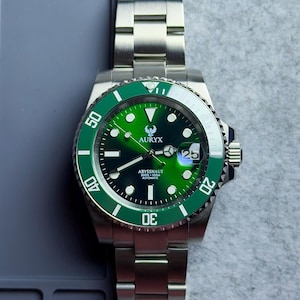 Op de afbeelding: Een roestvrijstalen polshorloge met een groene lunette en een groene wijzerplaat. De wijzerplaat toont de merknaam "AURYX" en de tekst "ABYSSNAUT 300m AUTOMATIC". Het horloge heeft een datumweergave op 3 uur en een roestvrijstalen band.