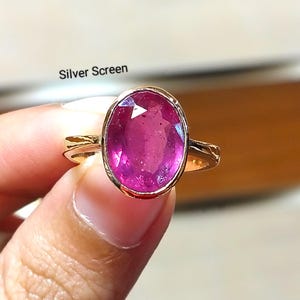 Pode incluir: Um anel de ouro com uma grande pedra preciosa rosa vibrante de corte oval, fixada num engaste liso. O aro do anel é fino e de metal dourado. O texto 'Silver Screen' está impresso em letras pretas pequenas acima da pedra preciosa.