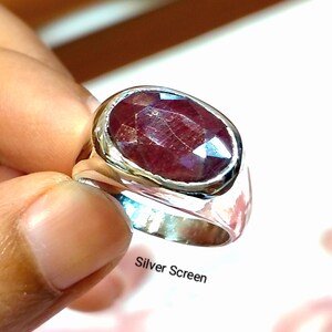 Puede incluir: Un anillo de plata con una gran piedra preciosa de color rojo oscuro, de forma ovalada y facetada, engastada en una banda de metal pulido. El anillo tiene una superficie lisa y reflectante con un engaste sólido que sujeta la piedra.