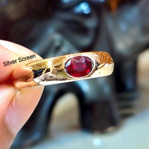 Peut inclure: Une bague en or avec une pierre précieuse rouge foncé de forme ovale sertie horizontalement au centre. La surface métallique est polie et réfléchissante, montrant des reflets lumineux. Le texte Silver Screen est imprimé près de l'anneau.